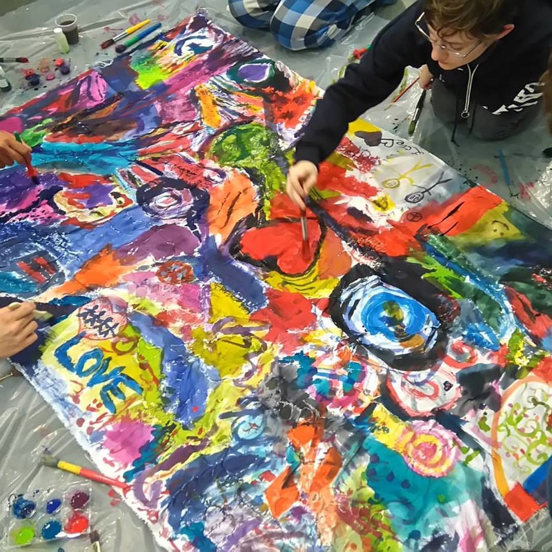 A teen wearing glasses and a black hoodie, lying on their stomach painting a colourful banner.