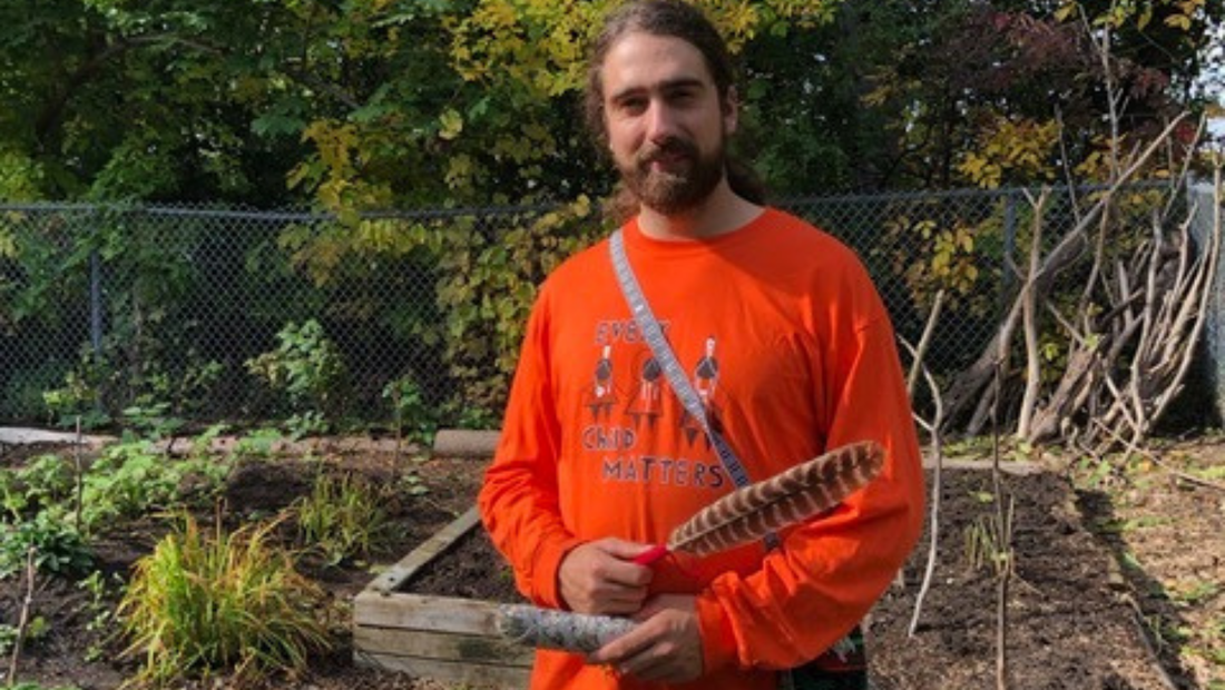 A bearded Indigenous man in an orange sweatshirt holds a sage wand and a feather for smudging. He is standing in the Ajashki Garden enclosure.