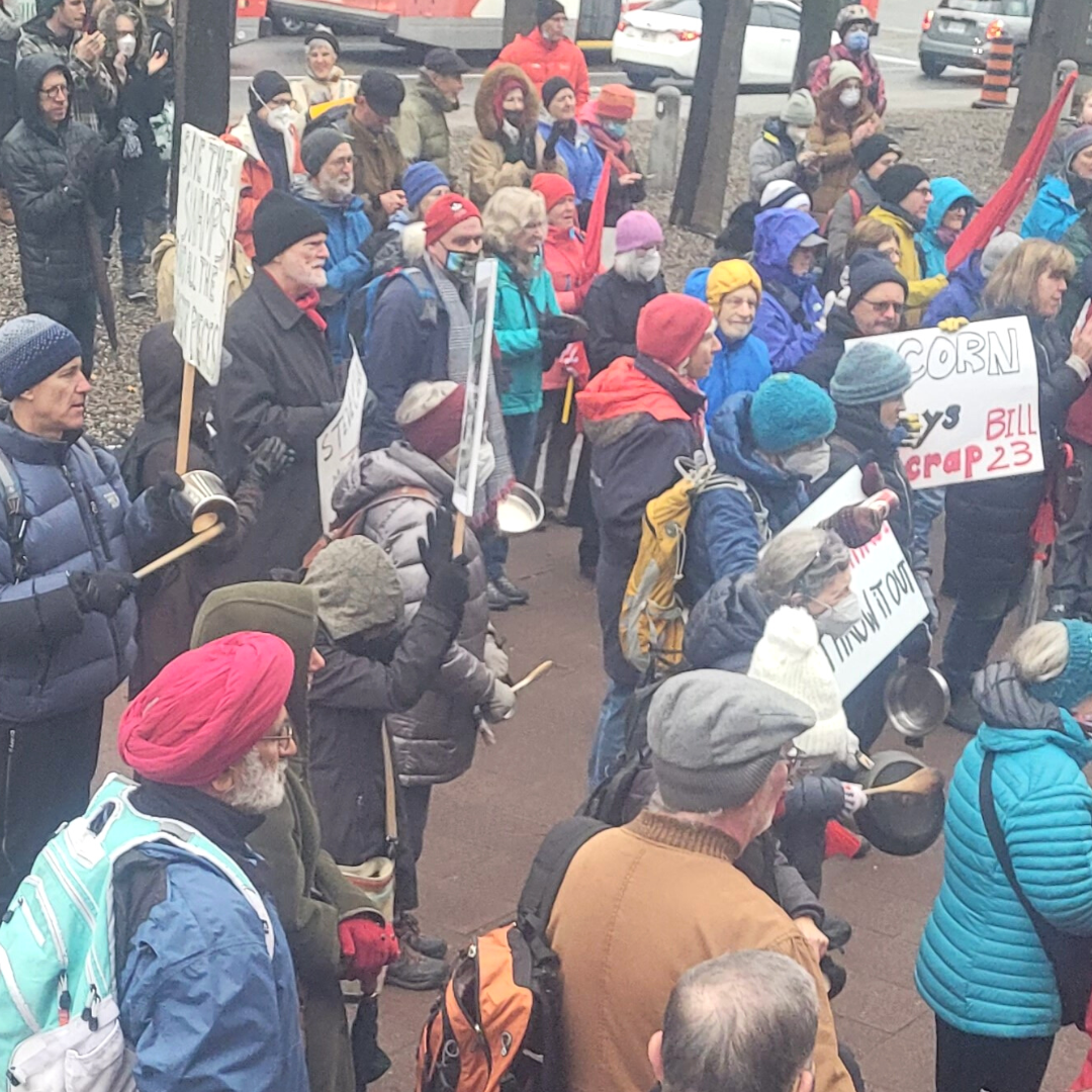 Another crowd shot featuring congregants wearing masks and winter coats. Some are carrying signs and some are banging on pots and pans.