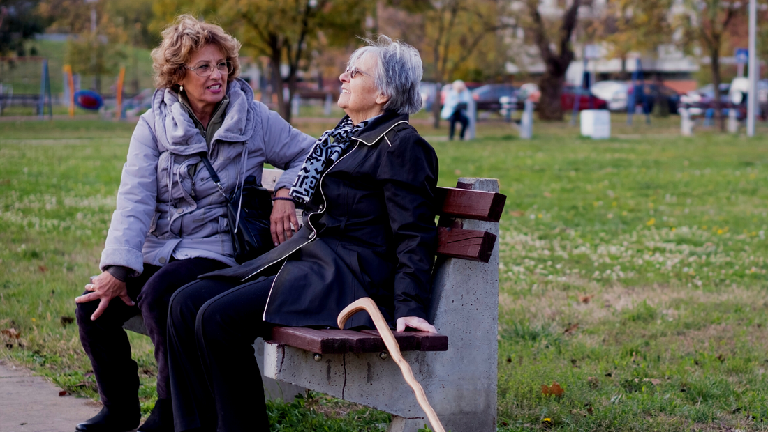 A middle-aged woman and an older woman with a white hair and a cane share a park bench together while they chat.