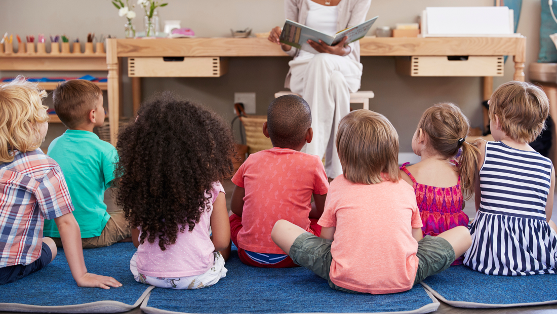 Seven little kids sit together on blue cushions, listening to a Black lady in white clothes read a story. There is a wooden work table in the background.