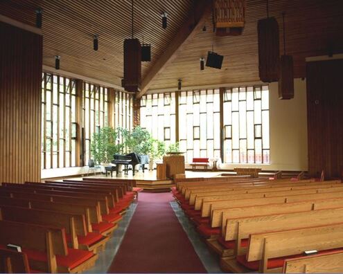 A photo of the FirstU Sanctuary, as viewed from the back of the room. The baby grand piano can be clearly seen at the front, in front of the wall of windows.