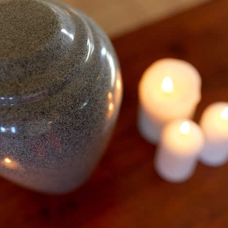 A grey stone funeral urn appears on a wooden table top next to three lit, white pillar candles.