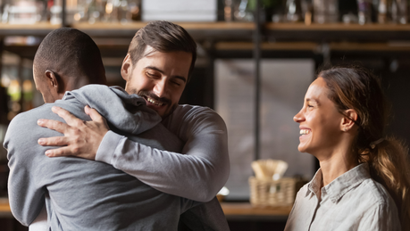 A Black man in a grey sweater is welcomed with hugs and smiles from his friends - a white man with short hair, and a woman in a dress shirt.