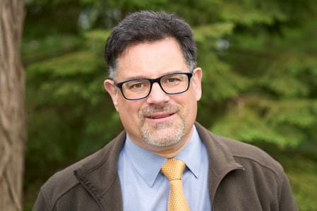 Reverend Eric - a middle-aged white man with a neatly trimmed beard - appears wearing a blue shirt and gold neck tie, as well as a casual brown jacket. He is pictured outdoors. He is the minister at the First Unitarian Congregation of Ottawa, a liberal, spiritual church located between Westboro and Britannia in Ottawa's west end.