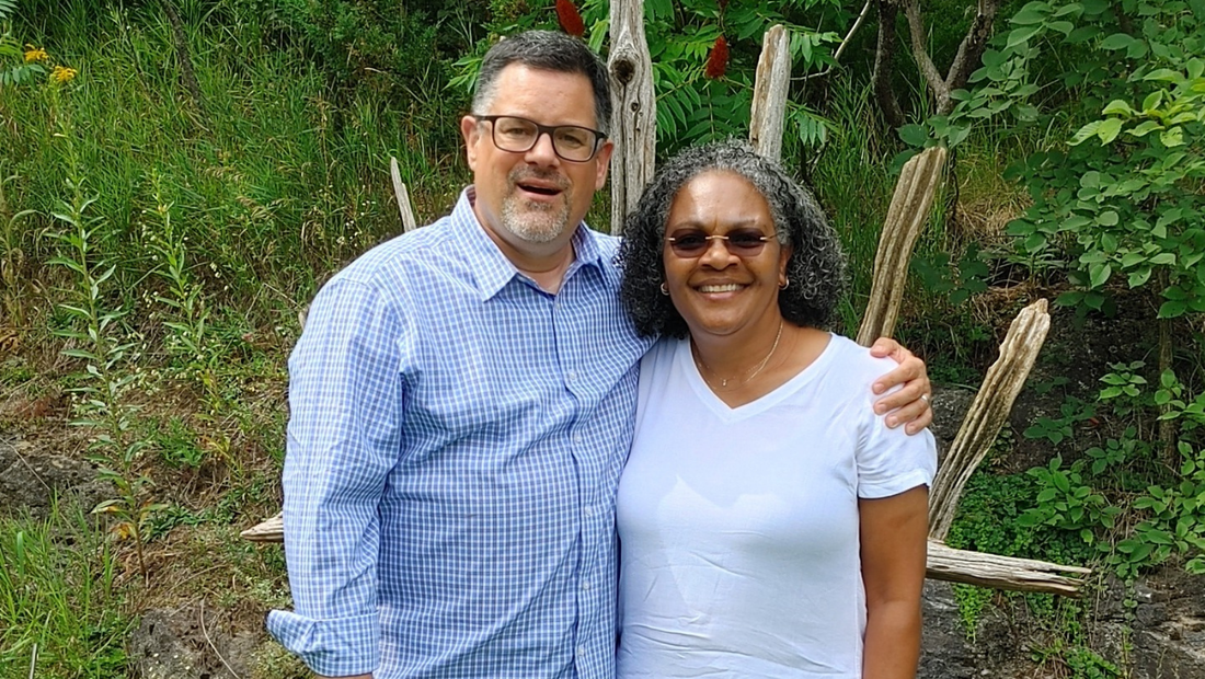 Reverend Eric (middle-aged white guy with a beard and a blue checked dress shirt) and Ann (middle-aged Black lady in a white t-shirt and sunglasses) stand, with their arms around each other, surrounded by greener. There is a rustic, wooden trellis behind them.