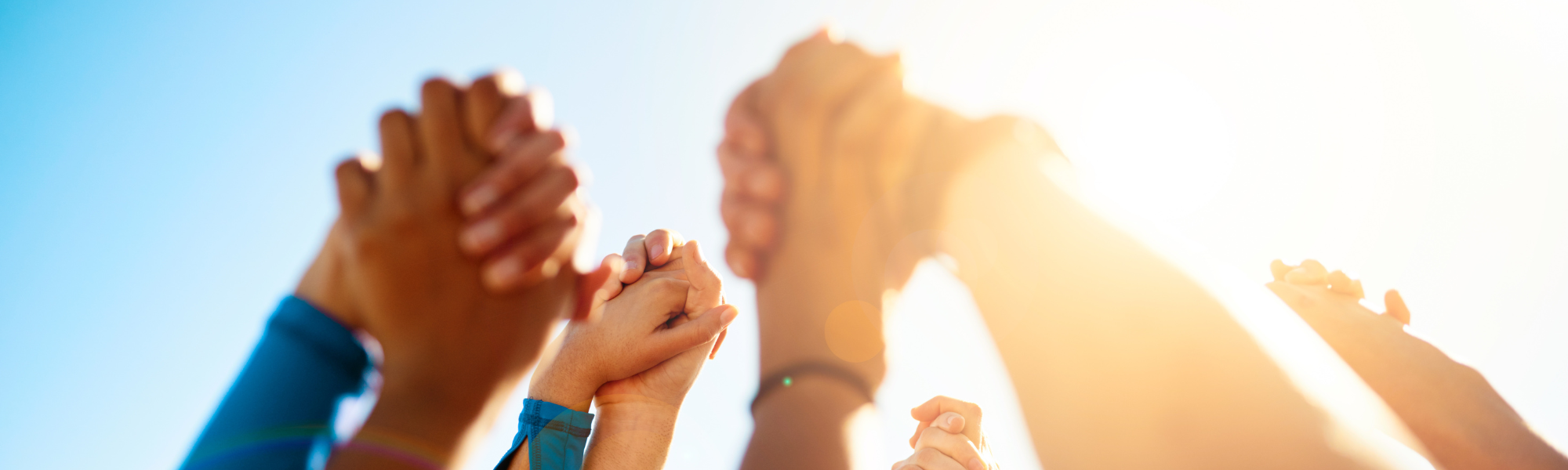 A stock photo of people's joined hands against a blue sky. The people are standing in a circle.