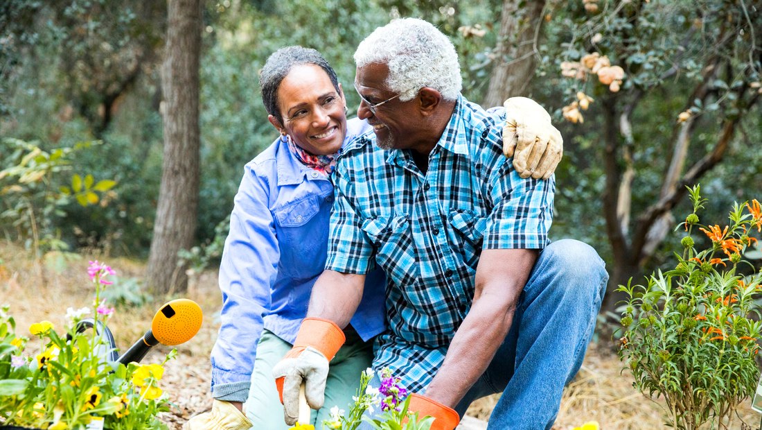 An elderly Black couple in casual clothes, working in the garden together. They are surrounded by the flowers and  young trees they've planted.