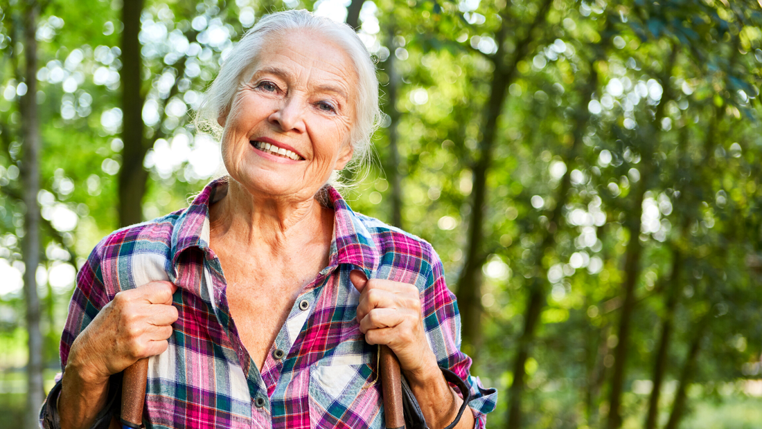 And elderly white woman in a plaid flannel shirt smiles at the camera. She is using two walking sticks as she makes her way along a woodland trail.
