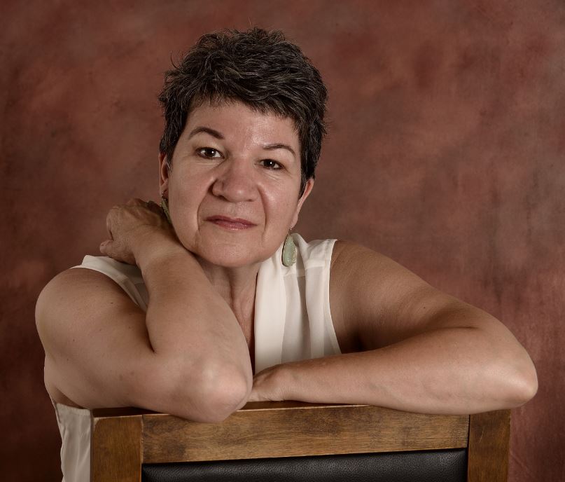 A headshot of Lia Boulay, a middle-aged woman in a white tank top and big earrings. She has a pixie haircut and is sitting backwards in a wooden chair.
