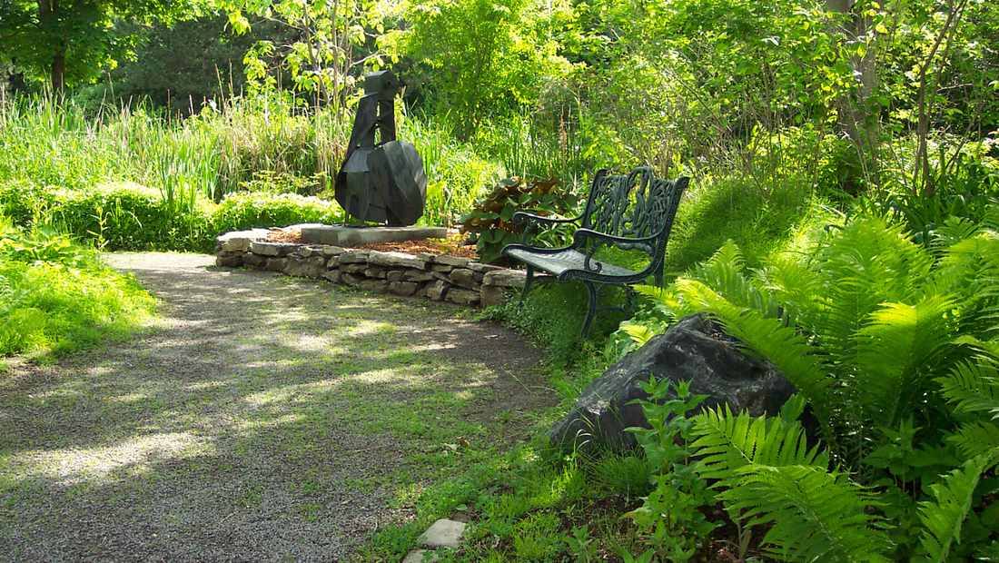 A photo of one of the wrought-iron benches situated in FirstU's Meditation Garden. It is located beside the walking path and surrounded by ferns and other summer greenery.