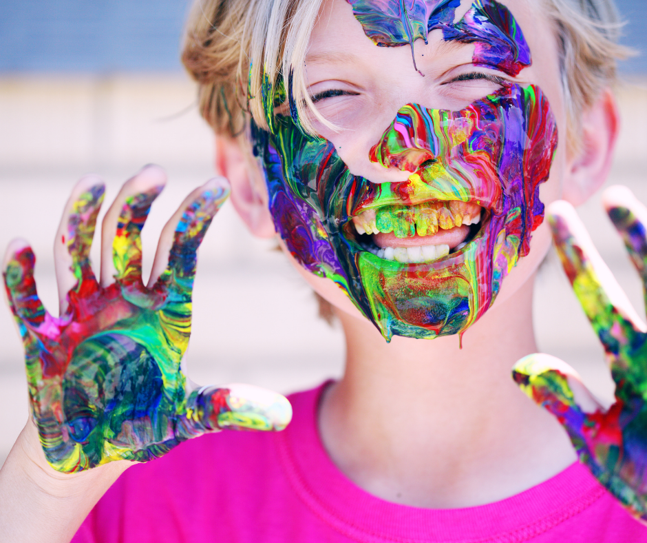 A grinning, blond child whose face and hands are covered with colourful paint. Photo by Sharon McCutcheon via Pexels.