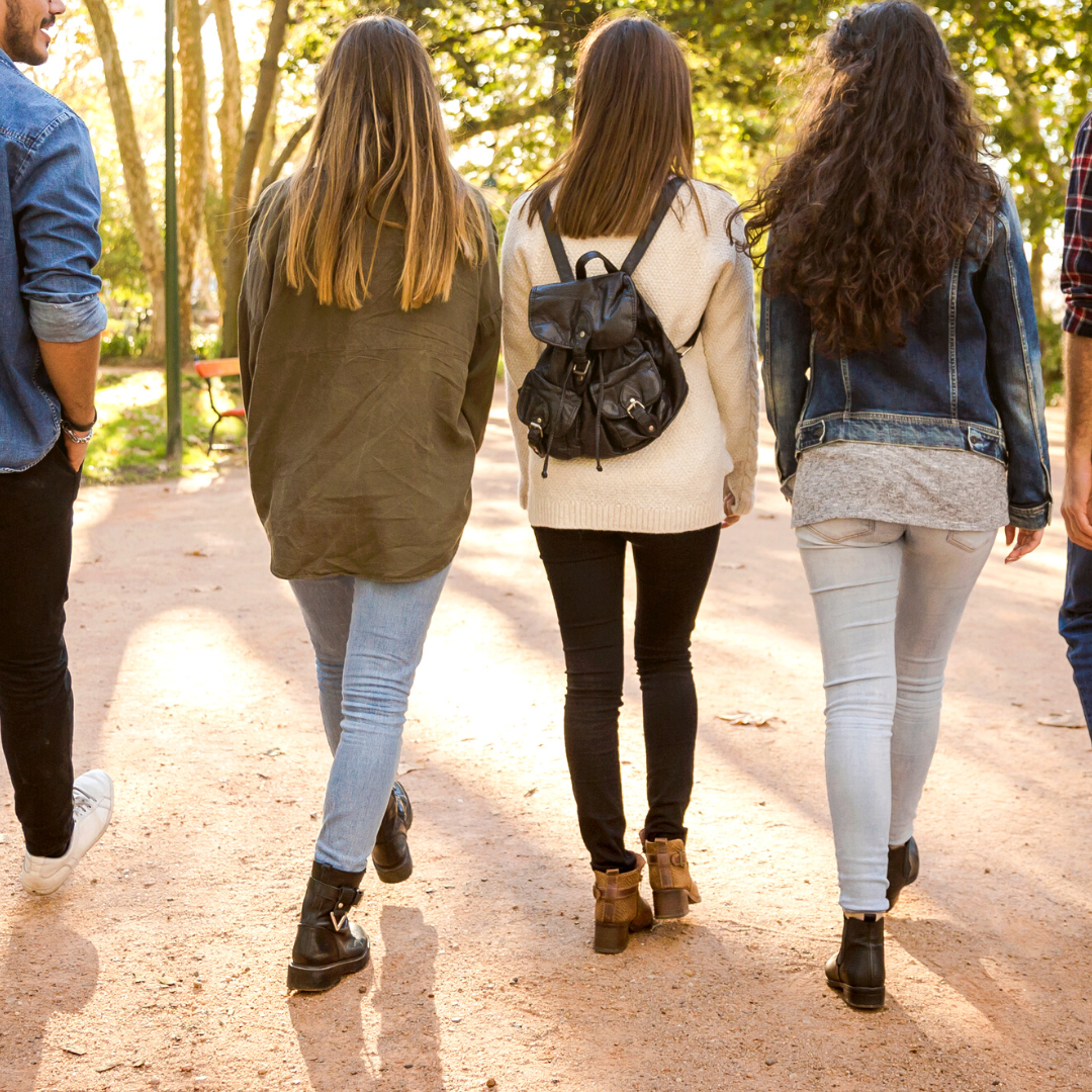 Four teens walking with an adult in their late 20s, as viewed from behind. They are all wearing casual clothes. There are trees in the background and they ware walking along a dirt path.